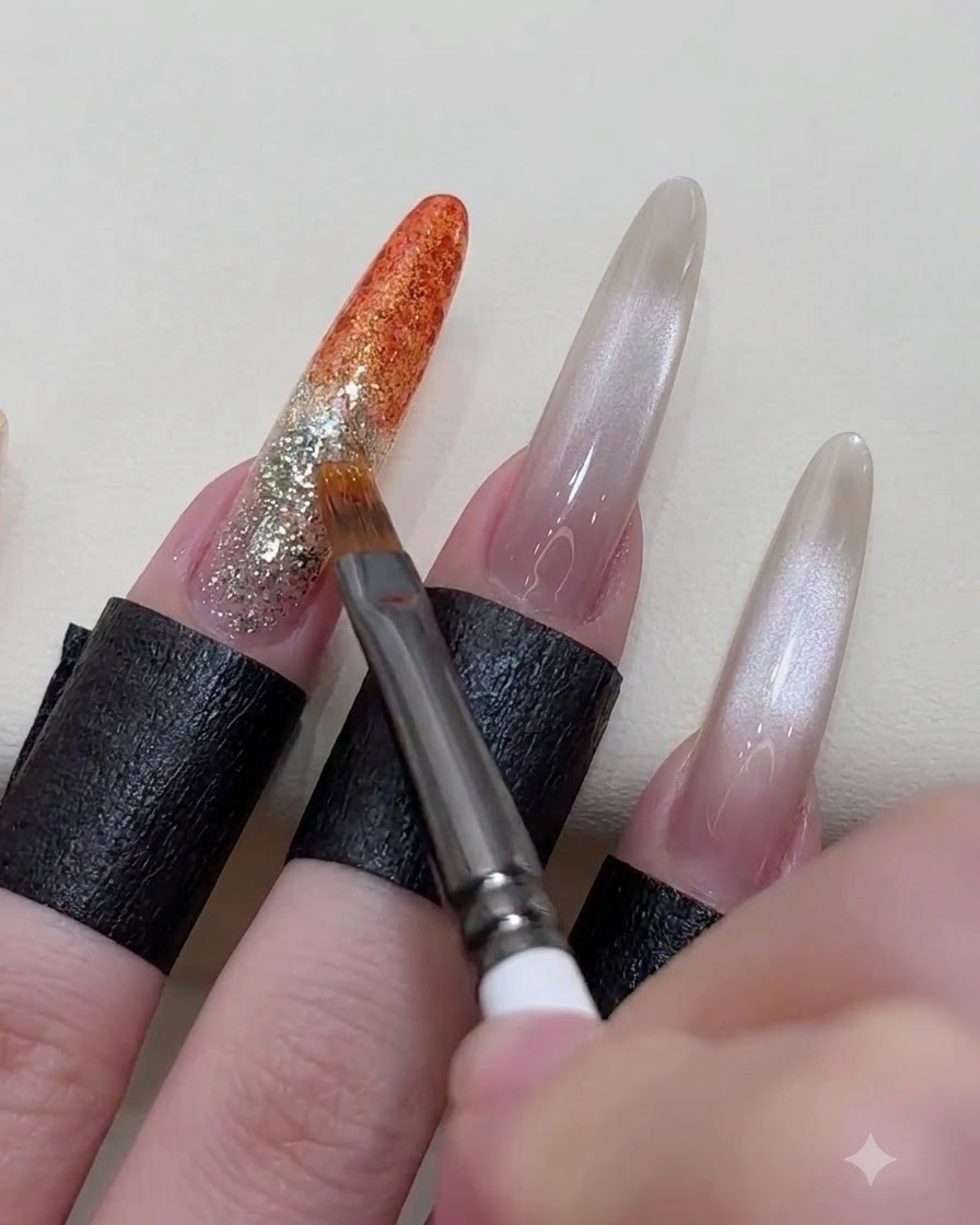 Eine Nahaufnahme einer Hand, die mit einem feinen Pinsel die Übergangslinie eines lebendigen Orangegels über einer Glitzer-Basisschicht verwischt und verblendet.