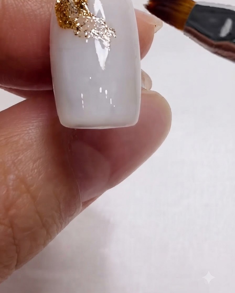 Ein Pinsel verblendet Goldglitter diagonal über die obere Ecke eines milchweißen Nagels.