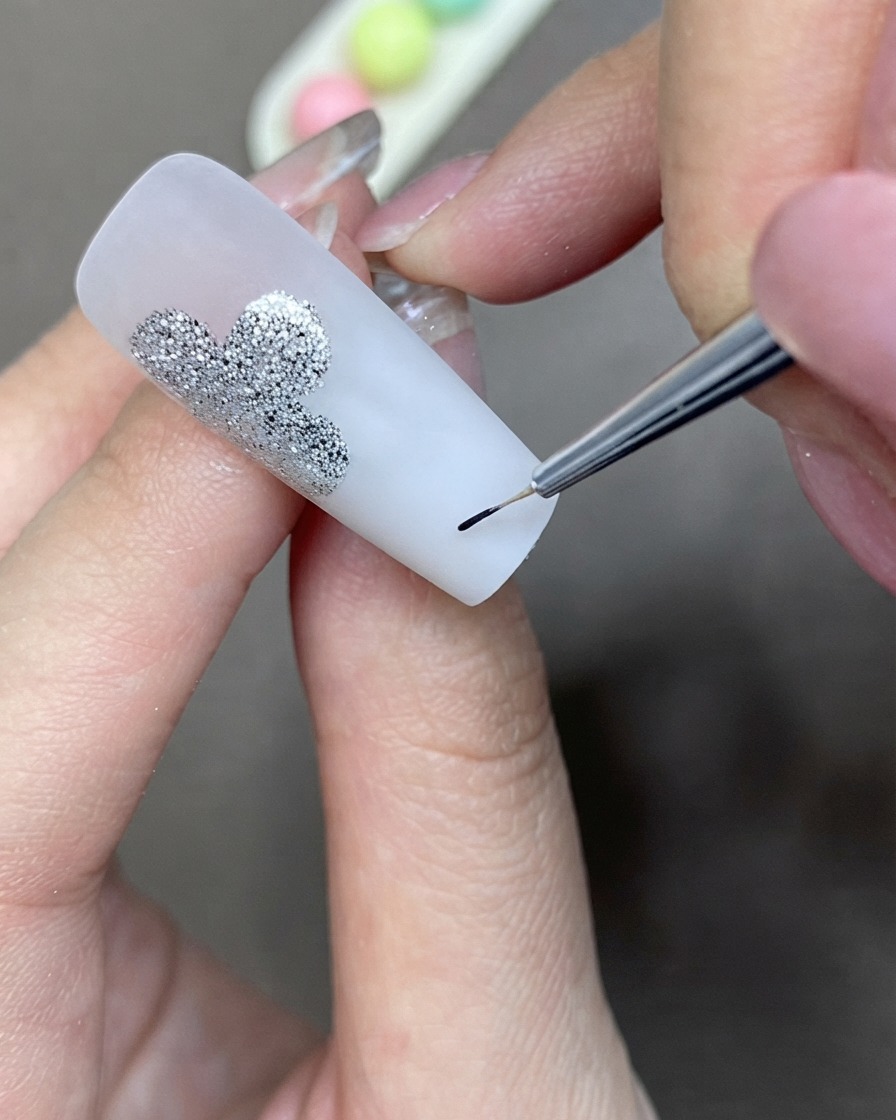 Ein feiner Detailpinsel trägt schwarzes Liner-Gel auf, um eine Kleeblattform neben einem silbernen Glitzerdesign zu kreieren.