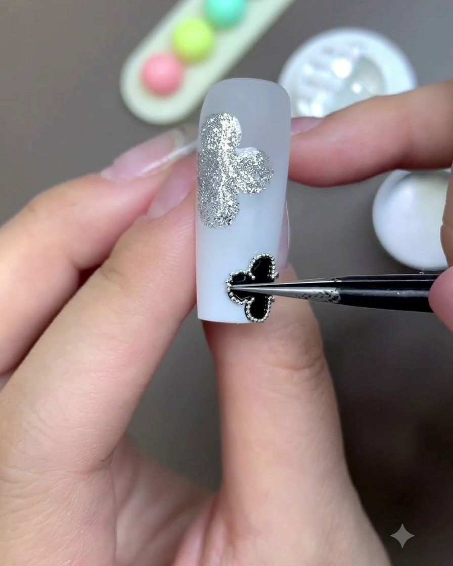 Eine Präzisionspinzette platziert ein gerahmtes schwarzes Kleeblattmotiv auf der Oberfläche eines milchig-weißen Nagels.