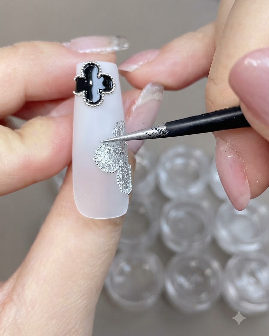 Ein feiner Pinsel platziert kleine Stahlperlen entlang des Randes eines silbernen Glitzer-Kleeblatts auf einem weißen Nagel.