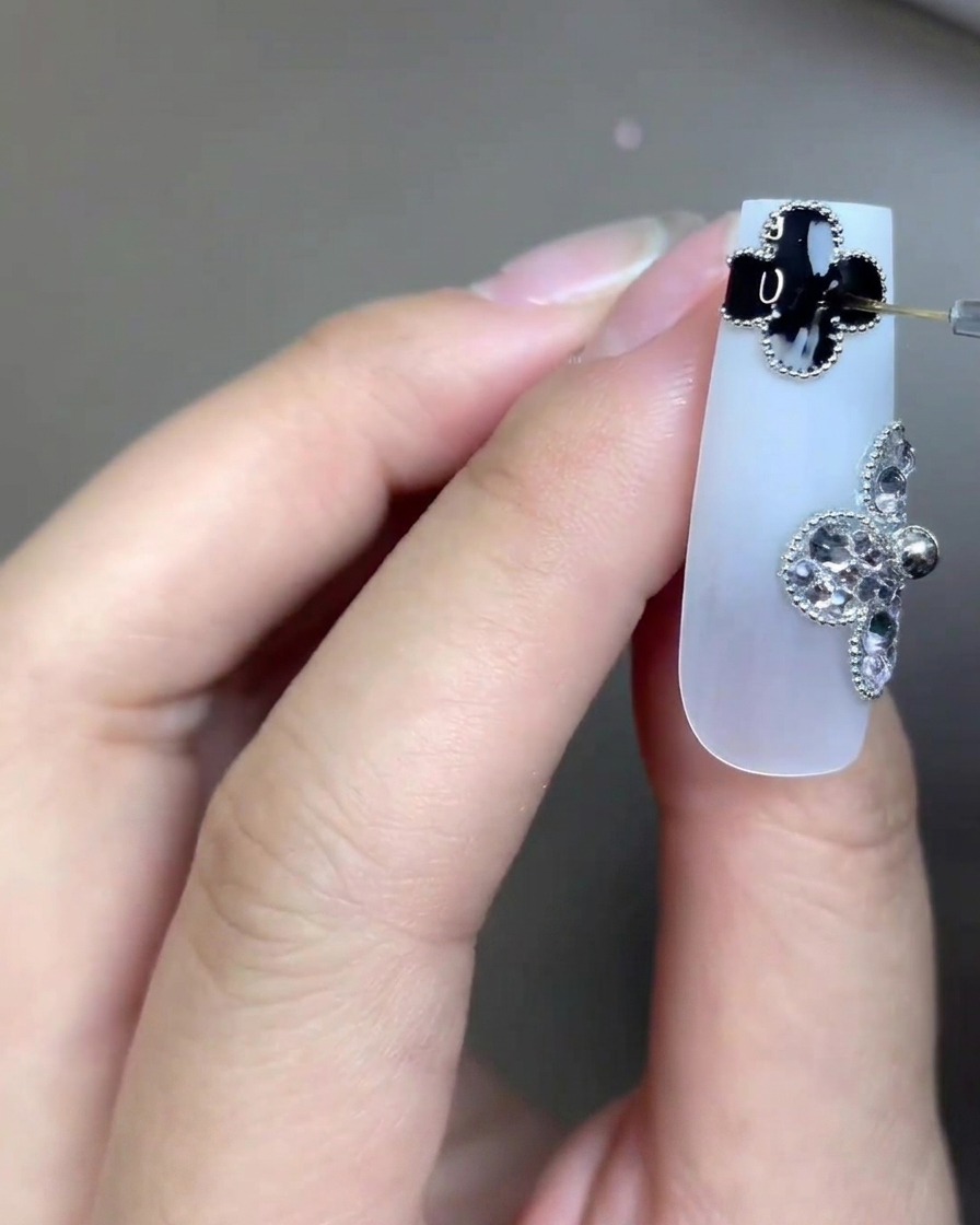 Nahaufnahme eines langen, milchig-weißen Nagels, verziert mit zwei vierblättrigen Kleeblatt-Anhängern – einer schwarz, einer aus Strasssteinen. Ein sehr feiner Nailart-Pinsel trägt präzise klaren Top Coat auf das schwarze Kleeblatt auf.