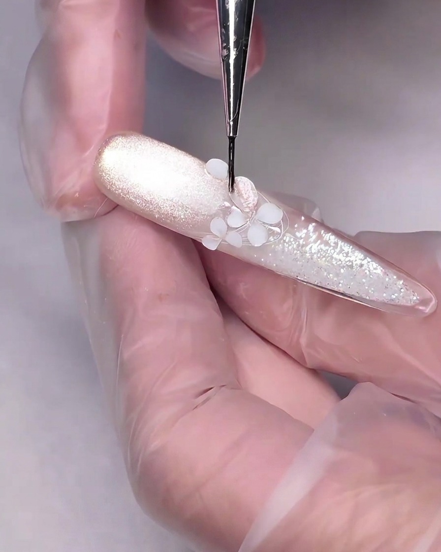 Ein feiner Detailpinsel trägt nudefarbenes Cat-Eye-Gel auf eine weiße, 3D-geschnitzte Blume auf einem Nagel auf.