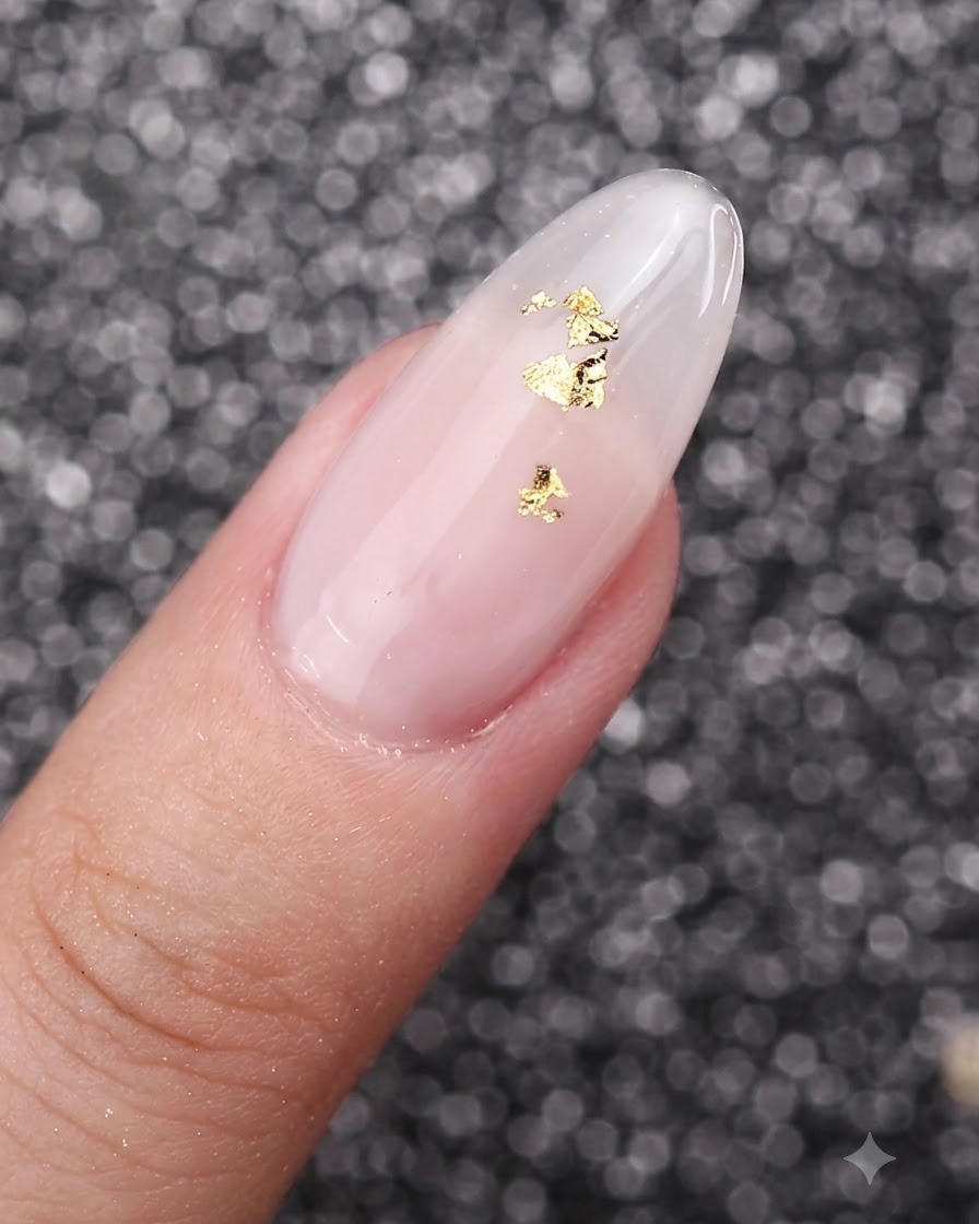 Eine Nahaufnahme von Blattgoldflocken, die zart auf einen mit milchig-weißem Gel lackierten Nagel gelegt werden.