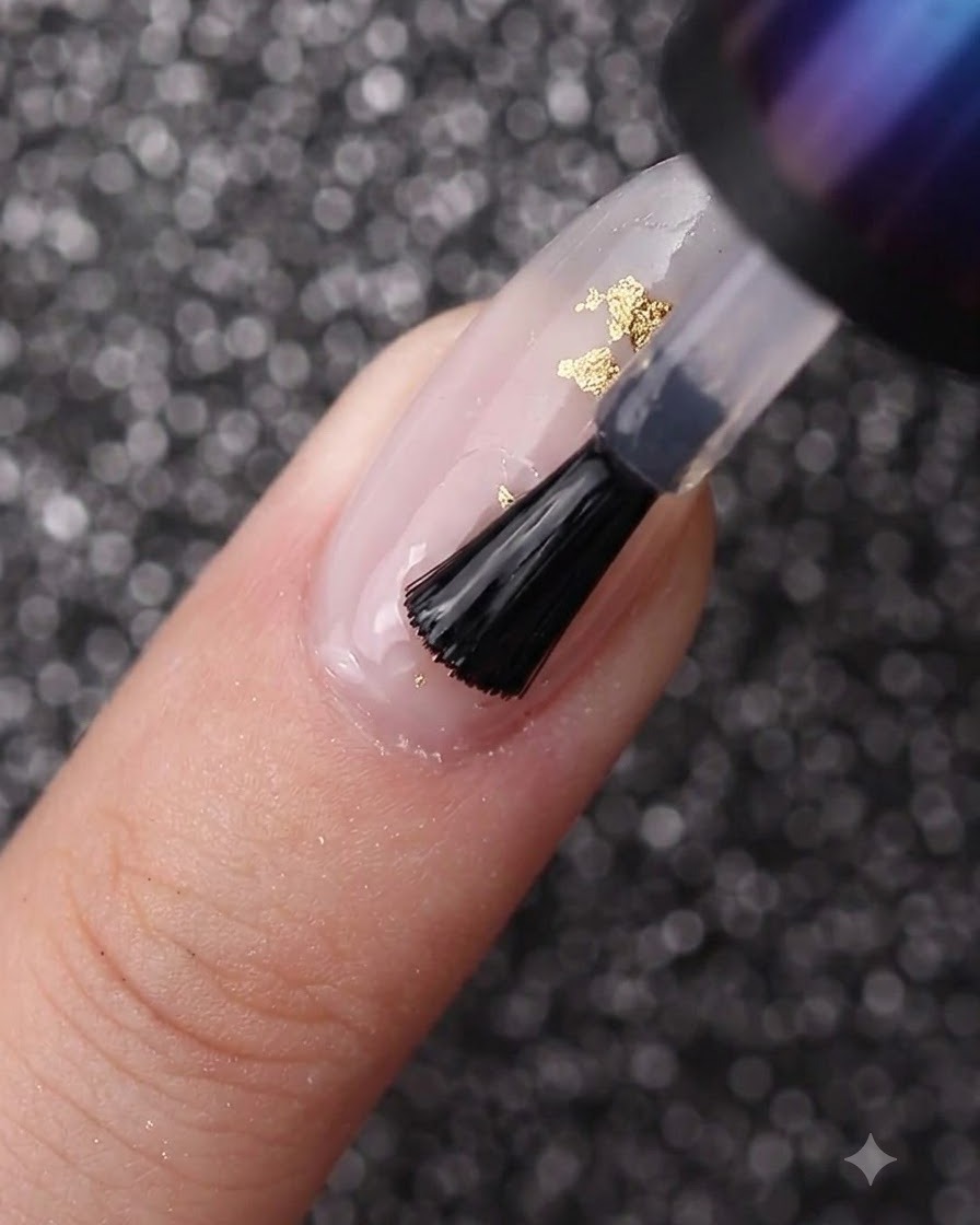 Ein Nagelpinsel verteilt klares Gel über einen mit milchig-weißem Lack und Blattgold verzierten Nagel.