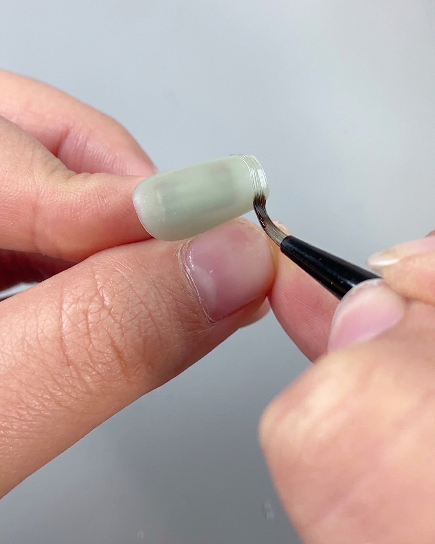 Ein feiner Detailpinsel verwendet klares Textur-Modelliergel, um eine erhabene Umrandung entlang der Kante eines Cat-Eye-Nagels zu ziehen.