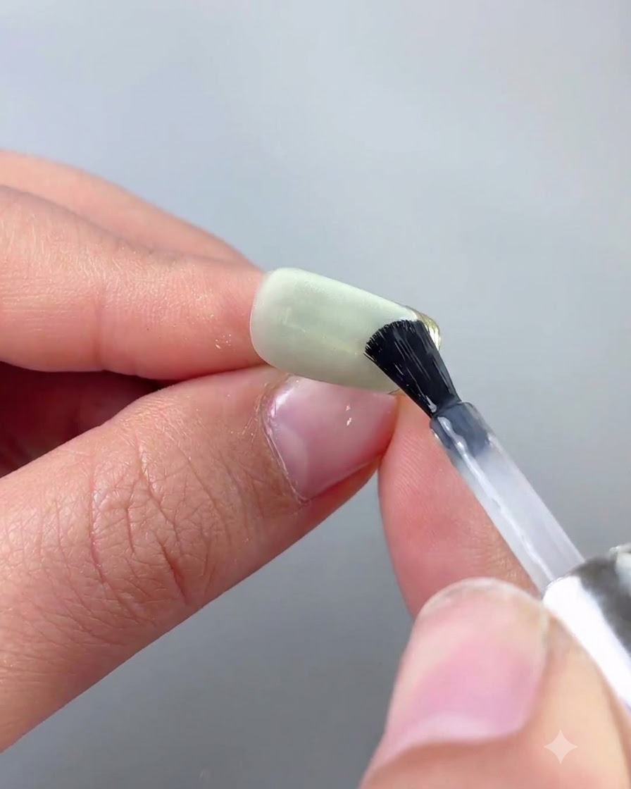 Ein feiner Nail-Art-Pinsel trägt Top-Coat präzise entlang einer Goldchromkante eines grünen Cat-Eye-Nagels auf.