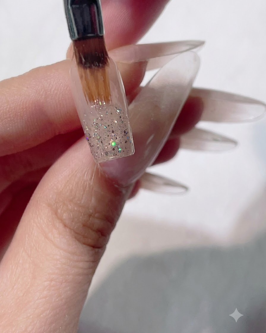 Ein flacher Gelpinsel streicht feuchtes Glitzer auf einem Nagel nach unten, um einen Ombré-Verlaufseffekt zu erzielen.