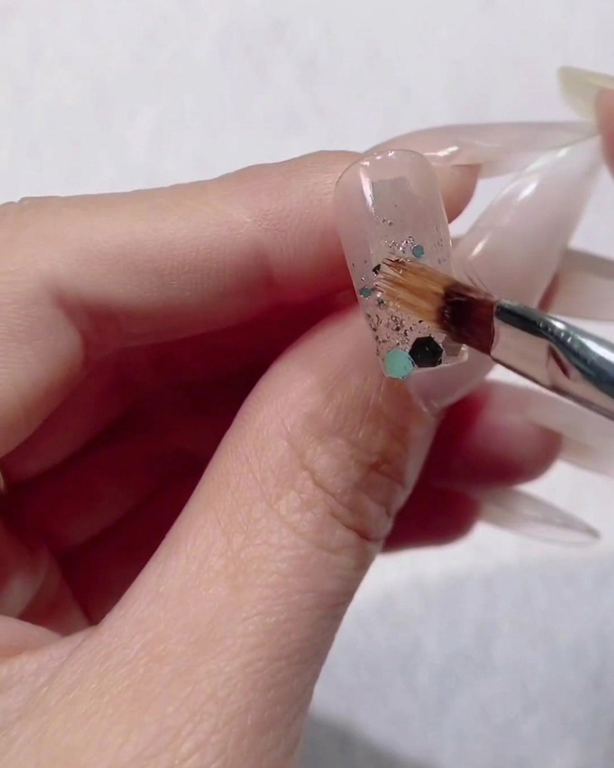 Ein kleiner Nagelpinsel platziert große blaue und grüne Sechseck-Pailletten auf dem feuchten Glitzerverlauf eines Fingers.