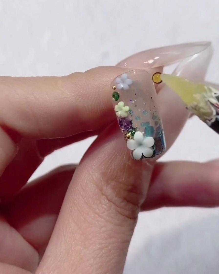 Eine Nahaufnahme einer Hand hält einen Fingernagel mit klarem Gel, Glitzer und winzigen 3D-Blumen, während ein Wachsstift einen gelben Strassstein platziert.