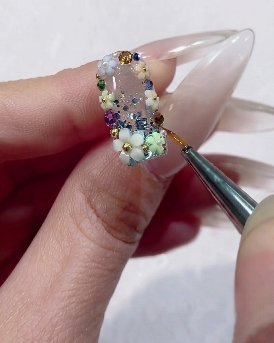 Ein feiner Liner-Pinsel trägt präzise Klargel um die Ränder eines facettierten Strasssteins auf einem Blumennageldesign auf.