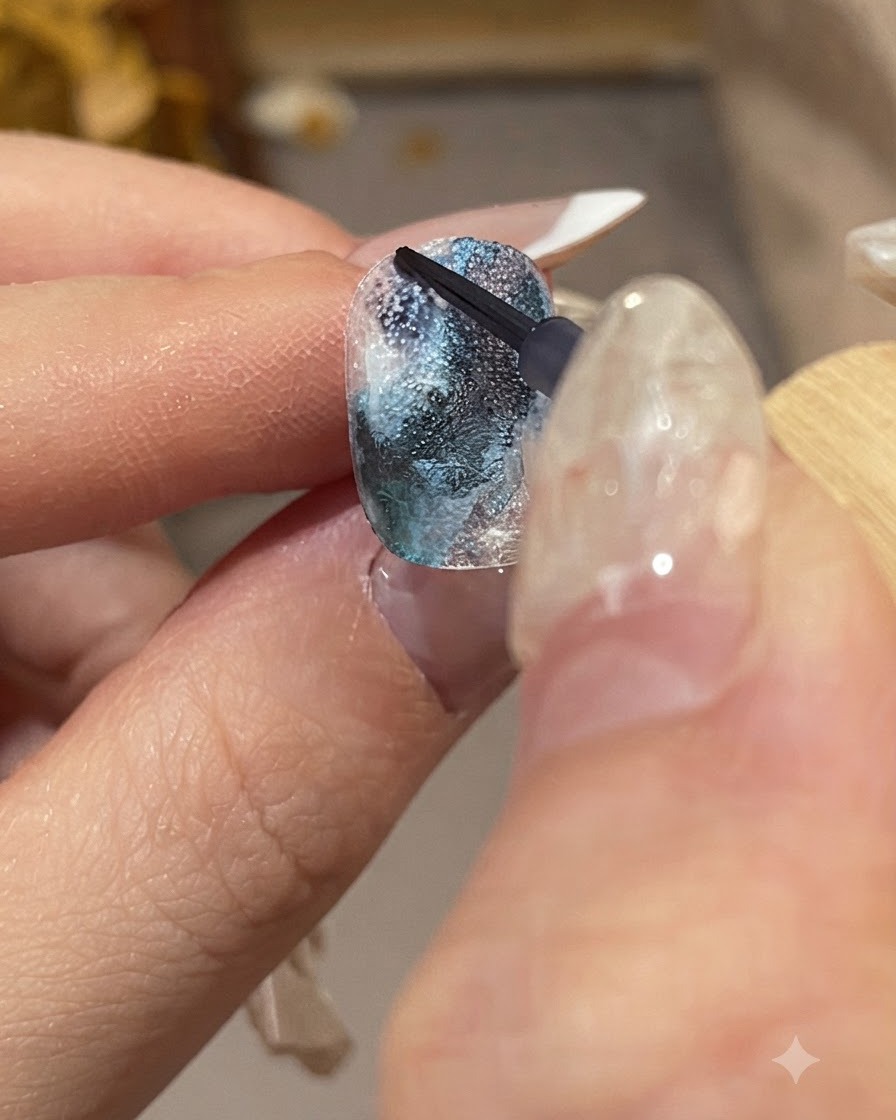 Eine detaillierte Nahaufnahme einer manicierten Hand, die einen feinen Pinsel hält, um dunkelblaue Tintentropfen auf eine strukturierte Nageloberfläche aufzutragen.