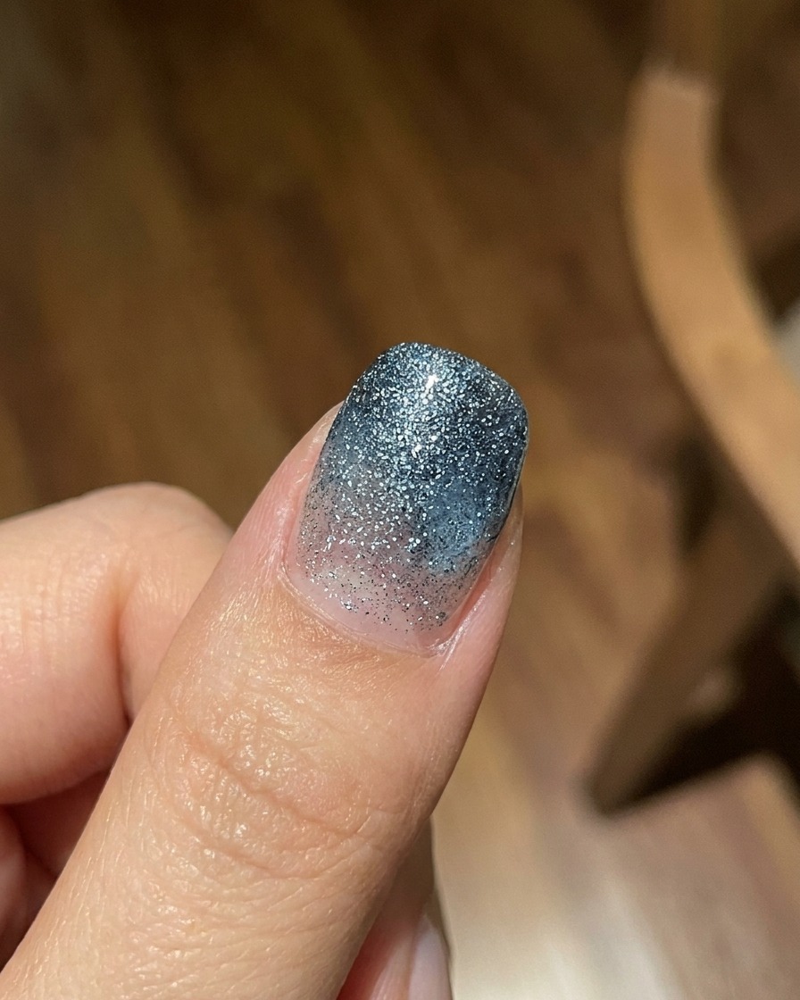 Ein Finger zeigt eine transparente blaue Jelly-Gel-Verlaufsschicht über einem strukturierten, glitzernden Hintergrund, aufgenommen in hellem natürlichen Licht.