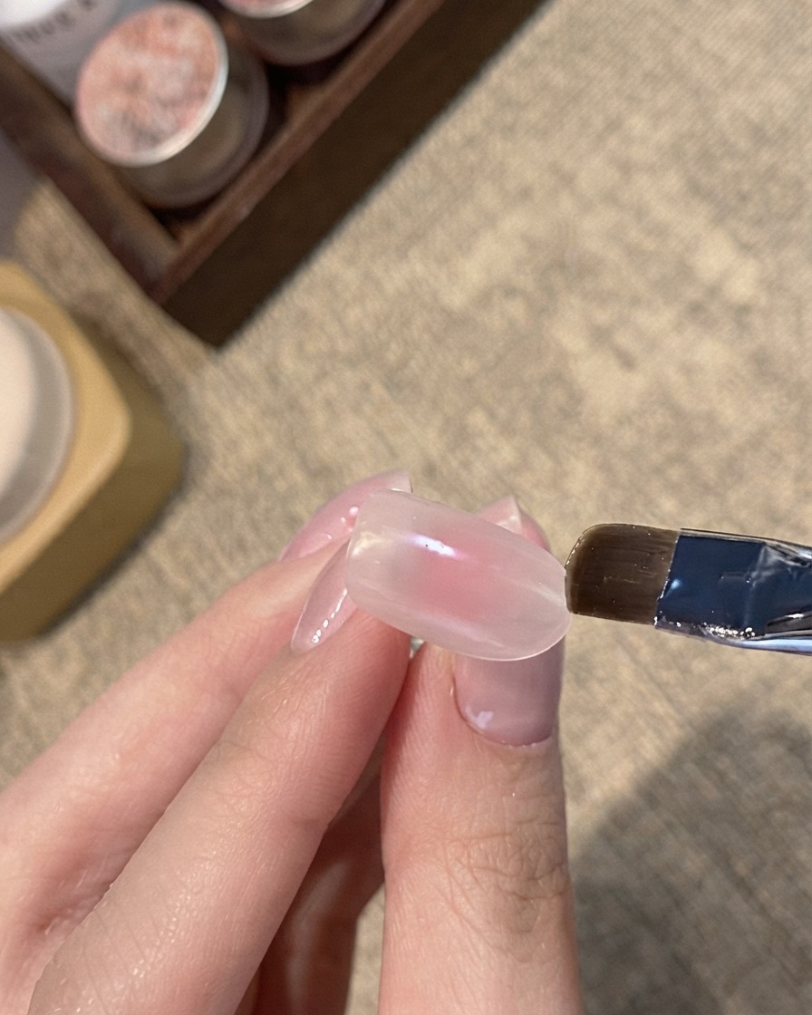 Ein flacher Pinsel trägt ein blaues Schimmer-Cat-Eye-Gel auf die Spitze eines rosafarben-nude Nagels auf.