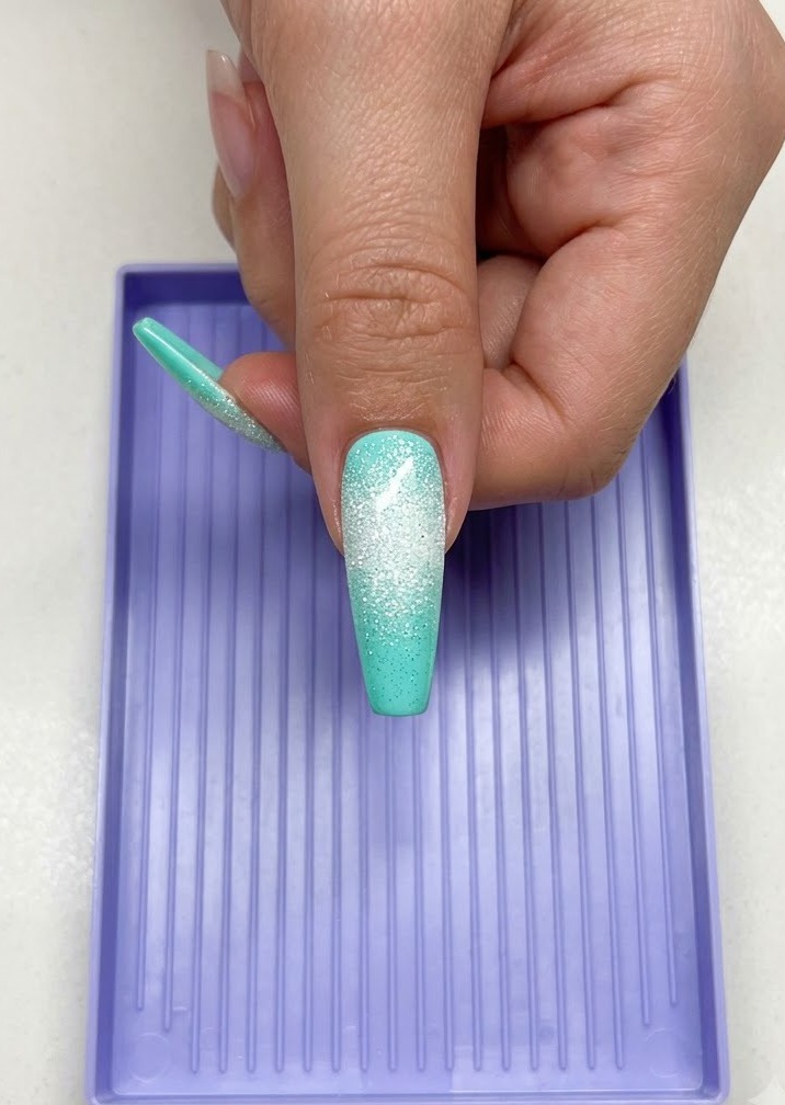 Eine Hand, die einen langen mintfarbenen Nagel mit einem weißen zuckerartigen Sterndesign unter einer lila UV-LED-Härtungslampe hält.