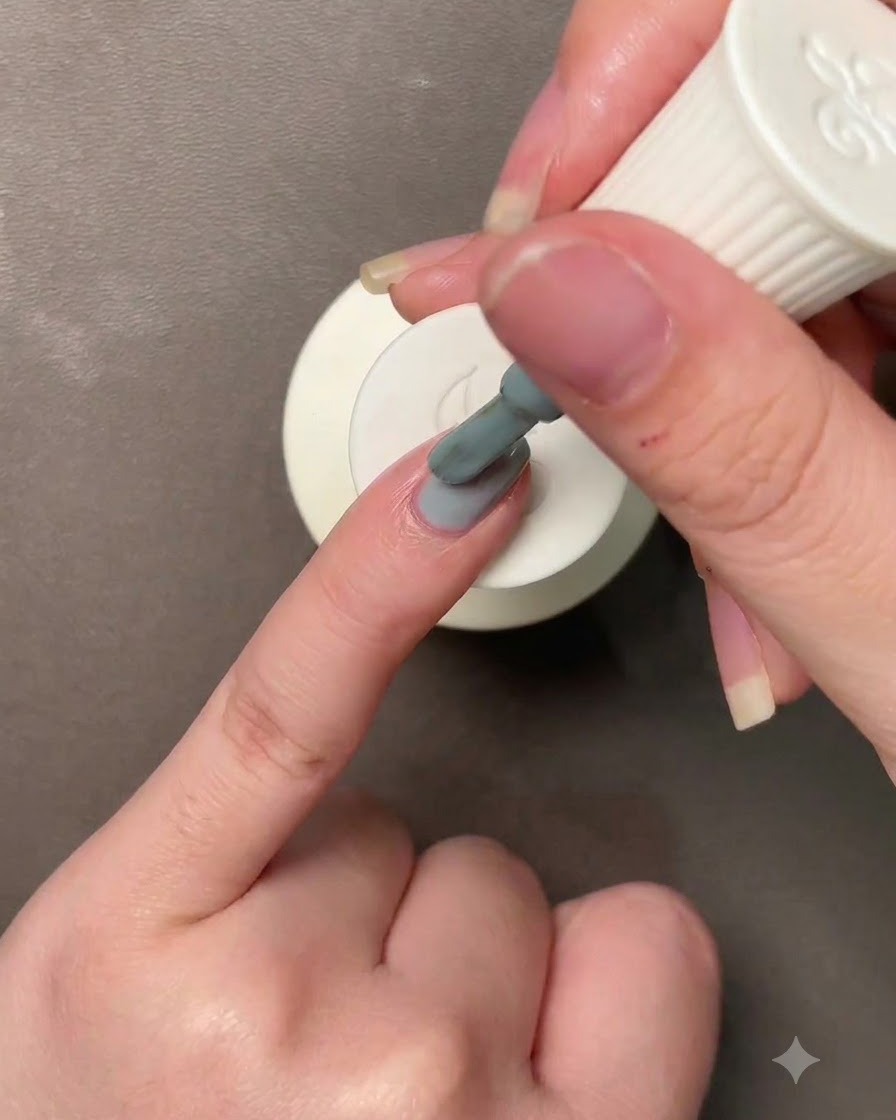 Eine Hand hält einen Pinsel und trägt graublau-grünes Gel auf einen Fingernagel auf.