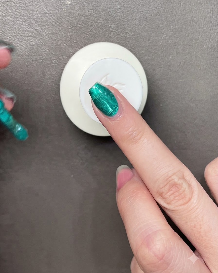 Ein Pinsel trägt lebendigen metallic-grünen Gel-Lack auf einen Nagel auf, während ein kleiner Magnet in der Nähe zu sehen ist.