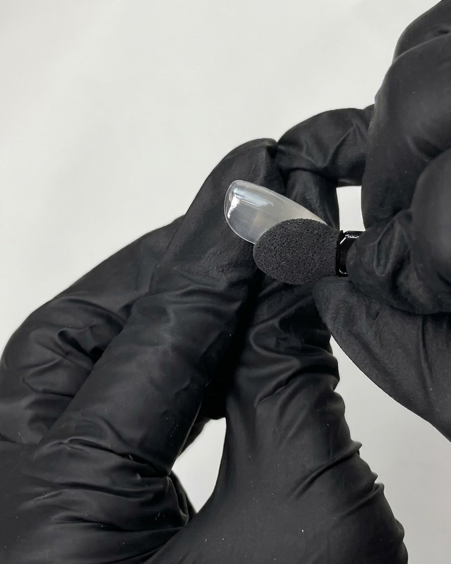 Eine schwarz behandschuhte Hand trägt mit einem Schwammapplikator irisierendes Chrompuder auf eine glatte künstliche Nagelspitze auf.