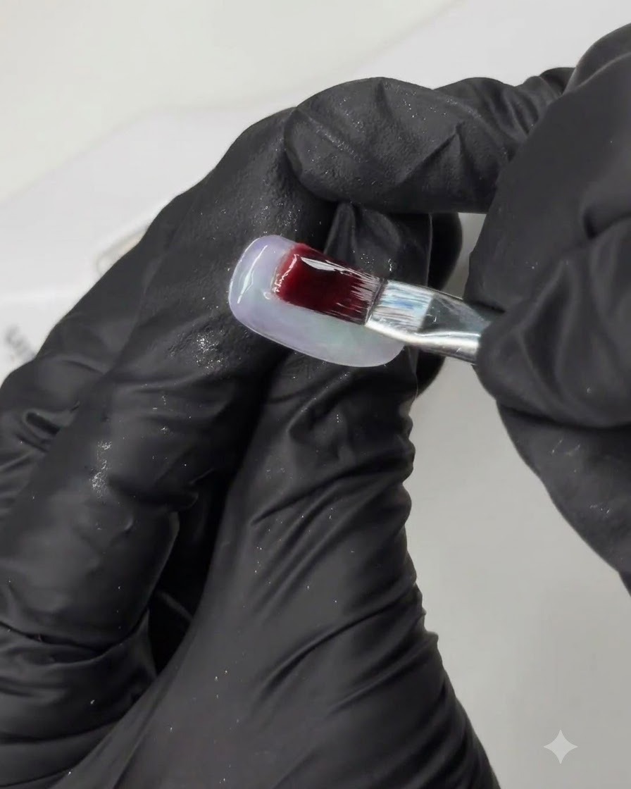 Ein Pinsel trägt ein tief pigmentiertes Gel während eines mehrstufigen Nagelkunst-Tutorials auf eine Übungsnagelspitze auf.