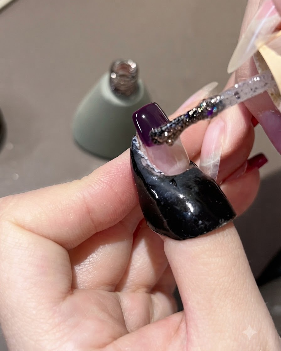 Ein Pinsel trägt lila Glitzergel über eine dunkellila Nagelspitze auf und zieht es leicht nach oben.