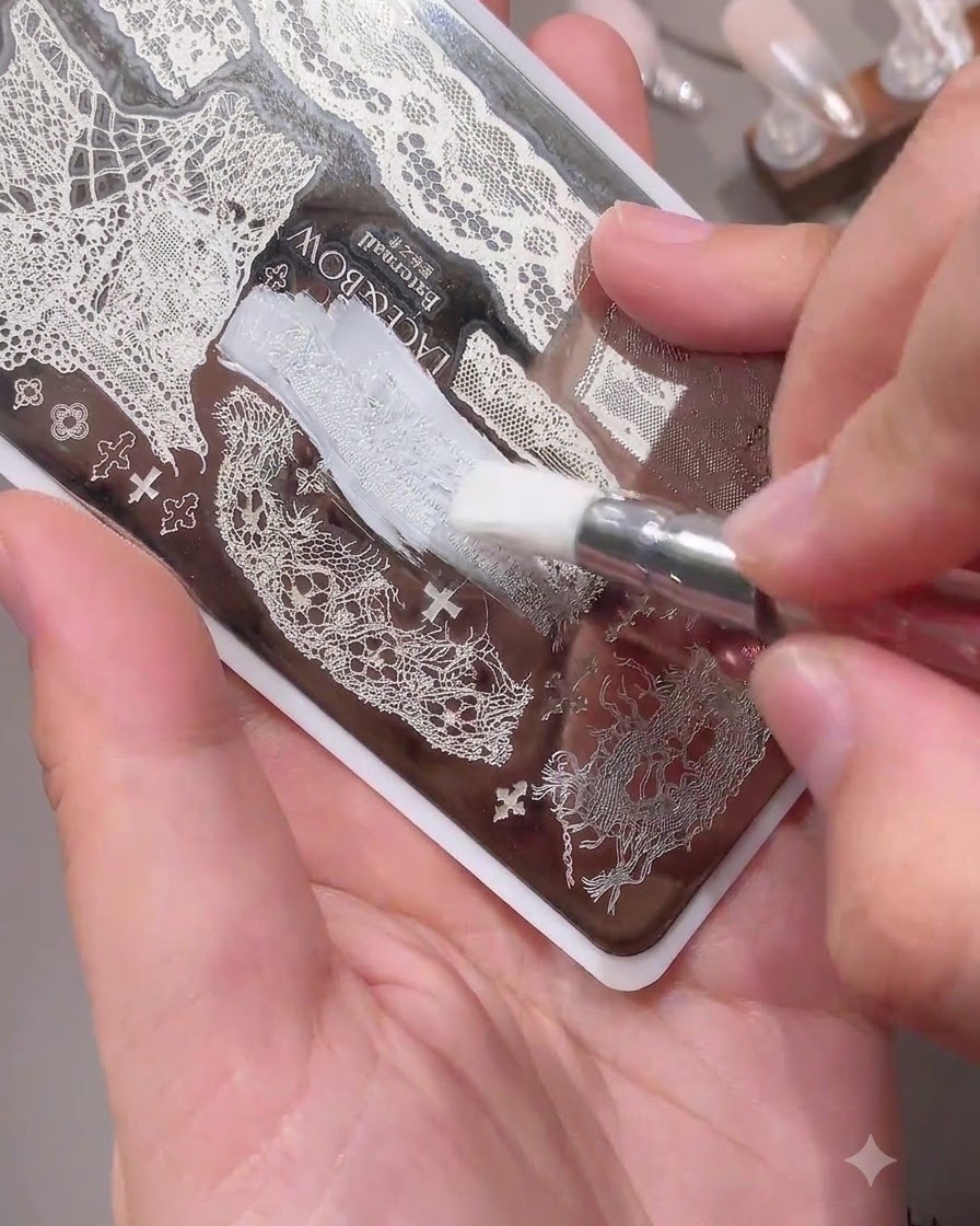 Ein Pinsel trägt weiße Farbe auf ein filigranes Spitzenmuster auf einer Nagel-Stamping-Platte aus Metall auf.