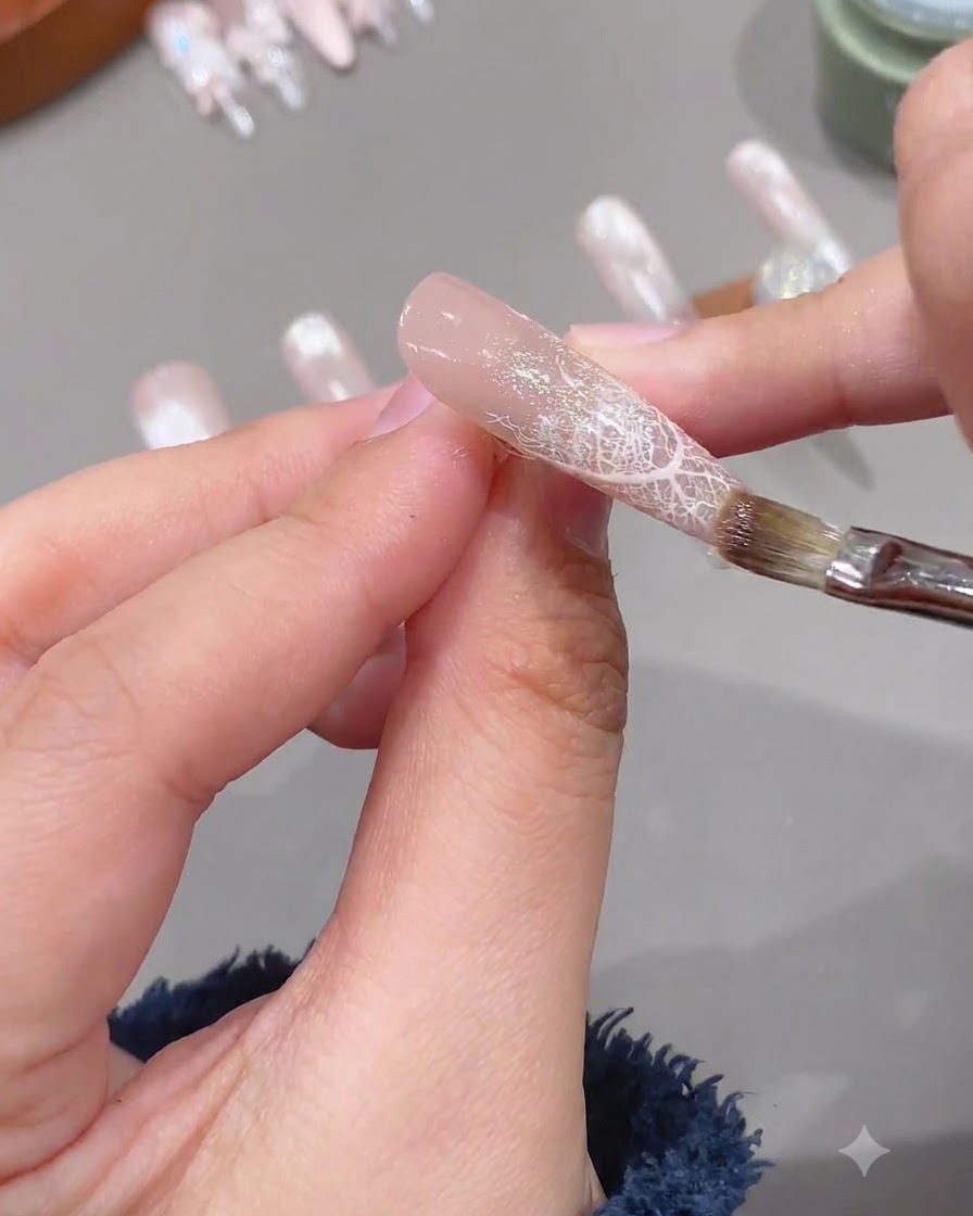 Ein Pinsel trägt eine klare Schicht weiches Verstärkungsgel auf einen vorbereiteten Nagel mit Spitzendesign auf.