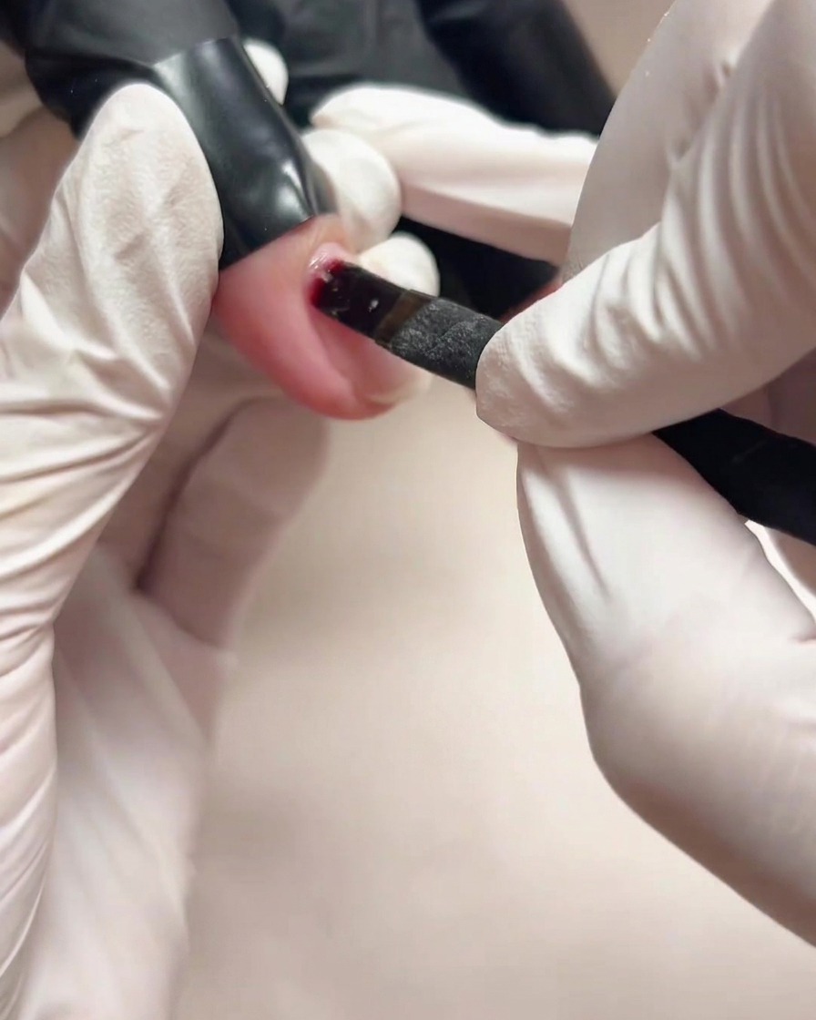 Nahaufnahme einer behandschuhten Hand, die mit einem kleinen Pinsel transparentes Base Gel auf einen Naturnagel aufträgt.