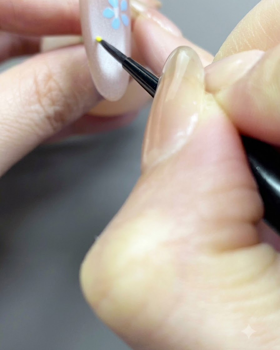 Ein feiner Pinsel detailiert einen kleinen gelben Mittelpunkt für ein florales Design auf einem Nude-Nagel.