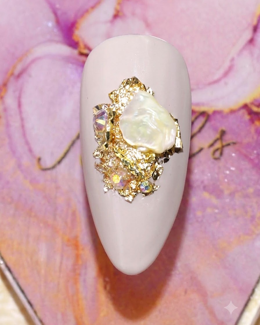 Eine Nahaufnahme eines verzierten Nagels mit einer Perlmutt-Muschel, Goldfolie und funkelnden Strasssteinen.