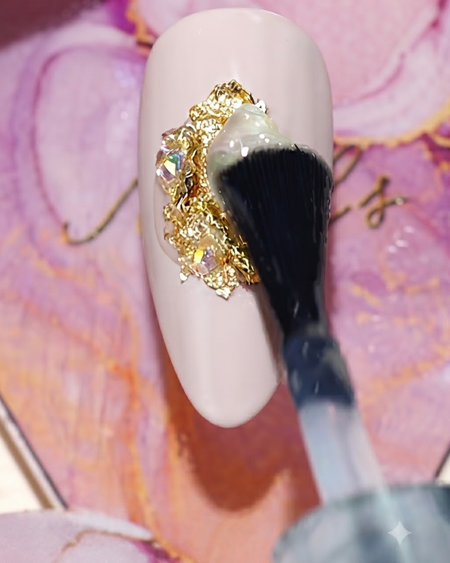 Ein Pinsel trägt einen klaren glänzenden Versiegelungs-Top-Coat über ein Cluster aus Goldfolie und Strass-Charms auf einem Nude-Nagel auf.