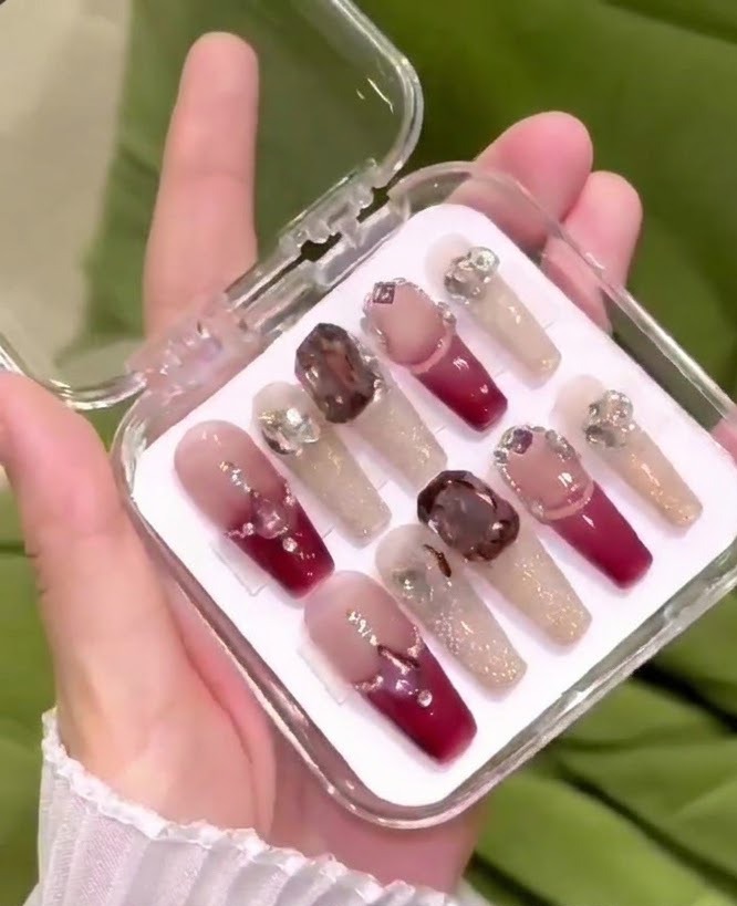 Eine Hand, die eine transparente Plastikaufbewahrungsbox hält, gefüllt mit Luxus Press-on Nägeln mit Rot-Nude-Verlauf und großen 3D-Kristallen.