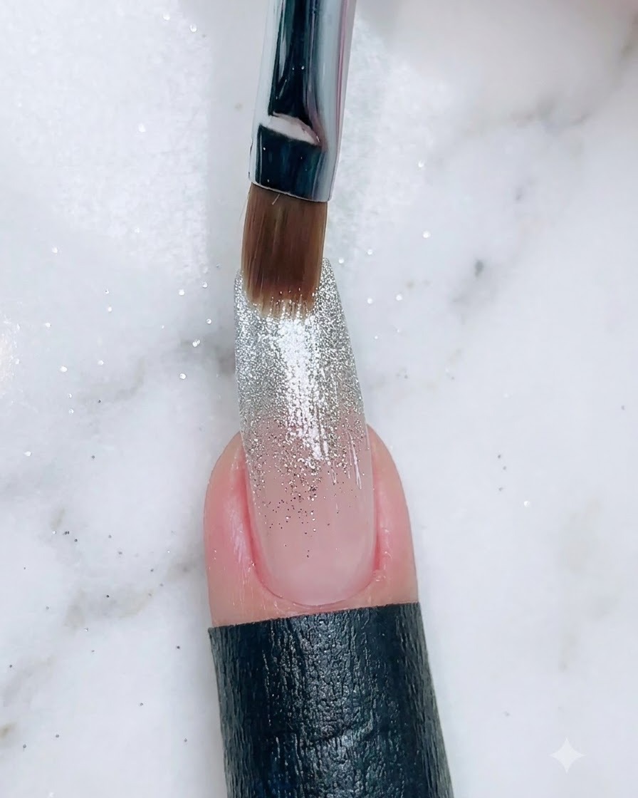 Ein Pinsel trägt eine dunklere Gelfarbe auf die Nagelbasis über einem Glitzer-Gradienten auf.