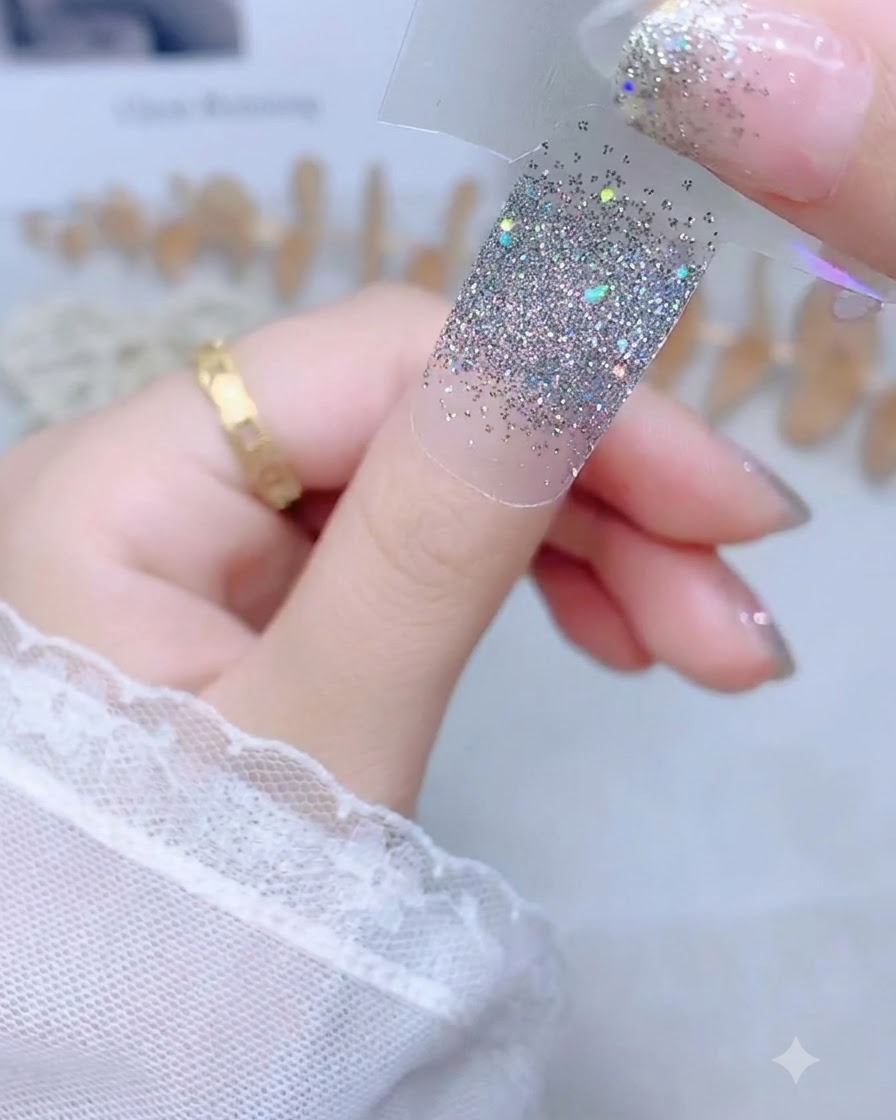 Eine Hand hält einen silbernen Glitzer-Nagelsticker an einem durchsichtigen Trägerstreifen über einen unlackierten Daumennagel.
