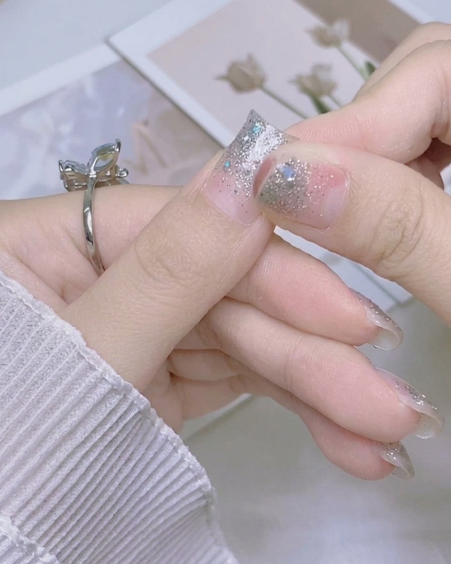 Eine Hand faltet die überschüssige Länge eines funkelnden, silbernen Nagelstickers nach unten über die Kante des Daumennagels.