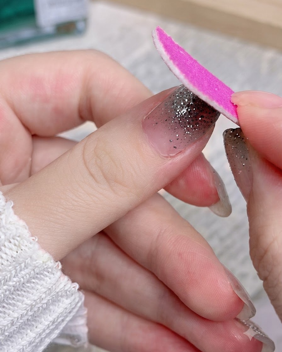 Eine rosa Nagelfeile feilt den überschüssigen Teil eines Silber-Glitter-Nagelstickers an einem Daumen ab.