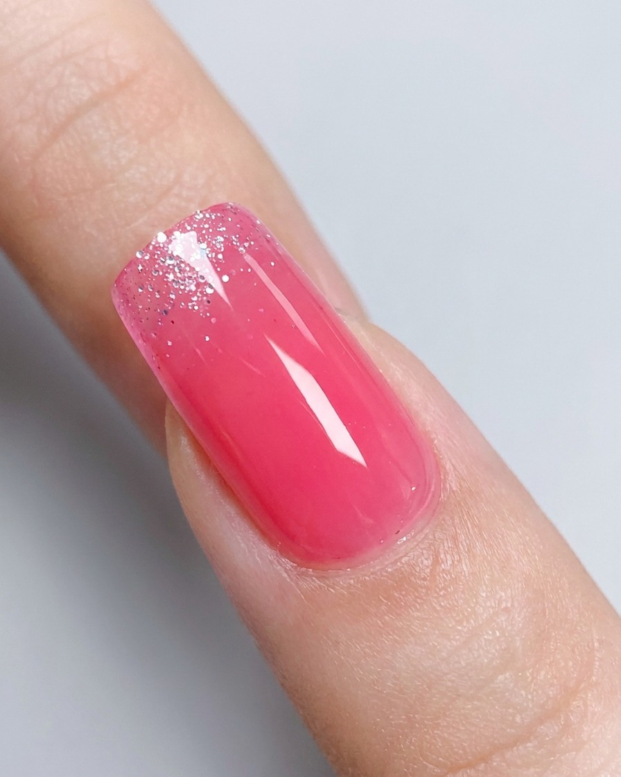 Eine Nahaufnahme eines glänzenden, rosa veränderten Fingernagels mit einem leicht verschwommenen Tiegel aus silbernem Glitter-Gel im Hintergrund.