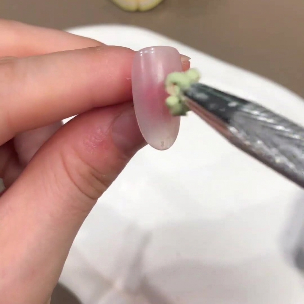Ein kleiner Pinsel trägt einen grün getönten Geltropfen auf die Mitte eines nude-rosa Nagels auf.