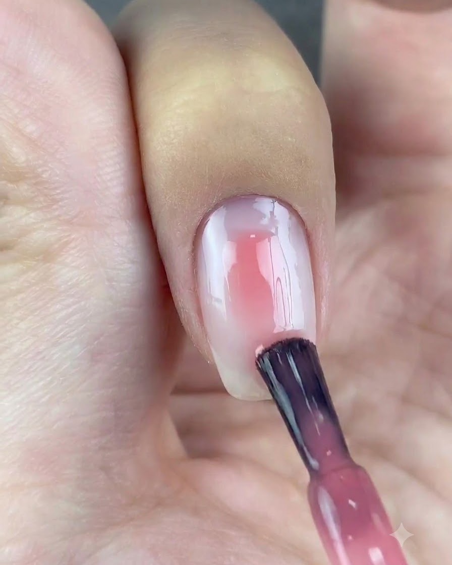 Ein Nagellackpinsel trägt einen Tropfen transluzentes Rosagel auf die Mitte des weiß grundierten Nagels auf.