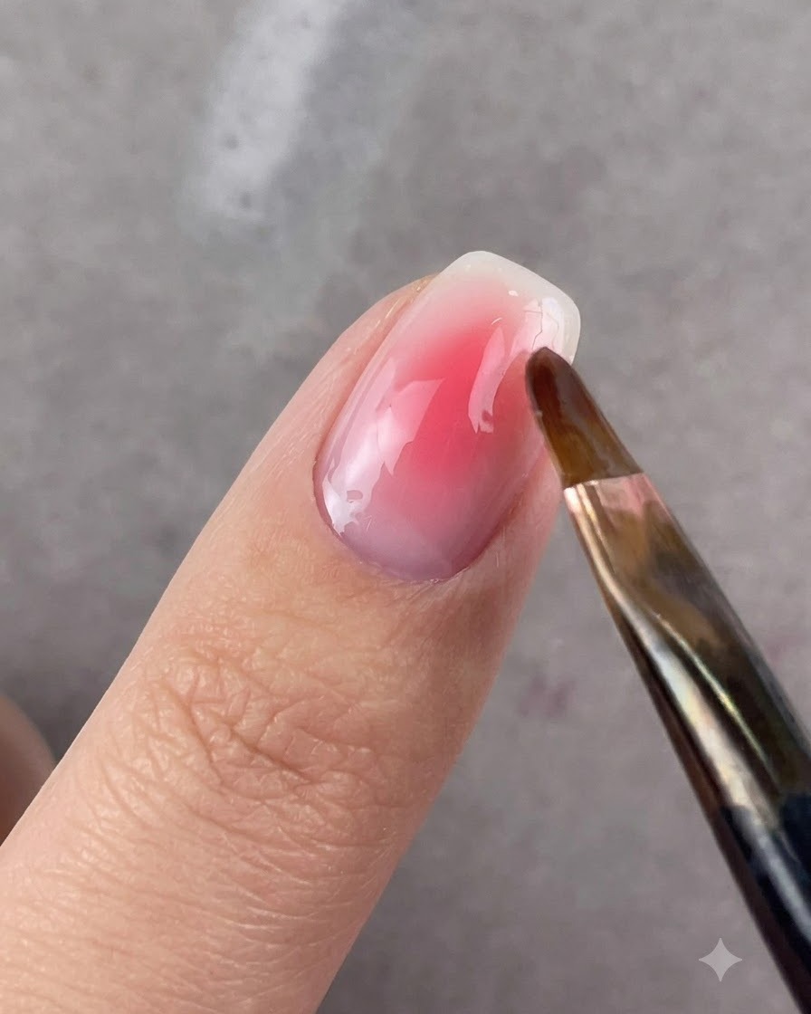 Ein runder Pinsel verblendet sanft die Ränder des Rosagels auf einem Fingernagel.