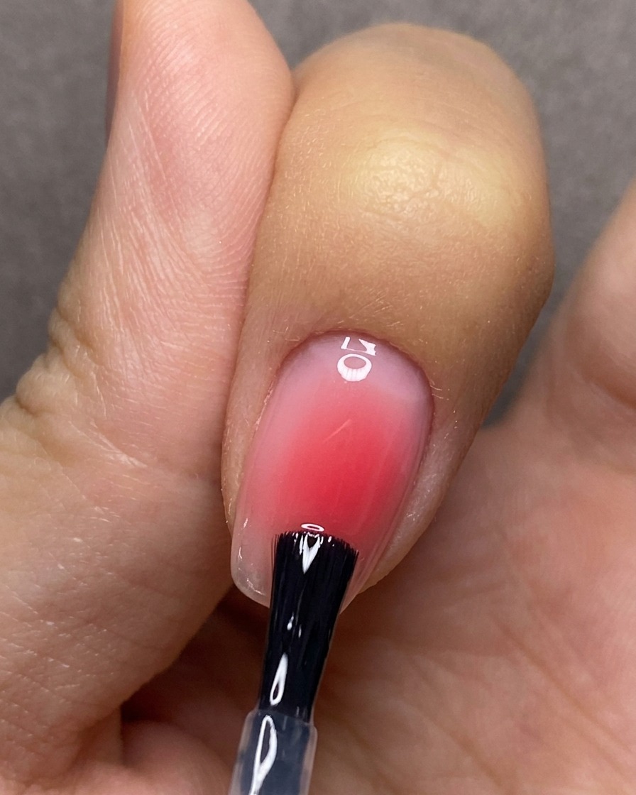 Ein Pinsel trägt einen klaren Top-Coat gleichmäßig über einen Nagel mit rotem Blush-Gradienten-Design auf.