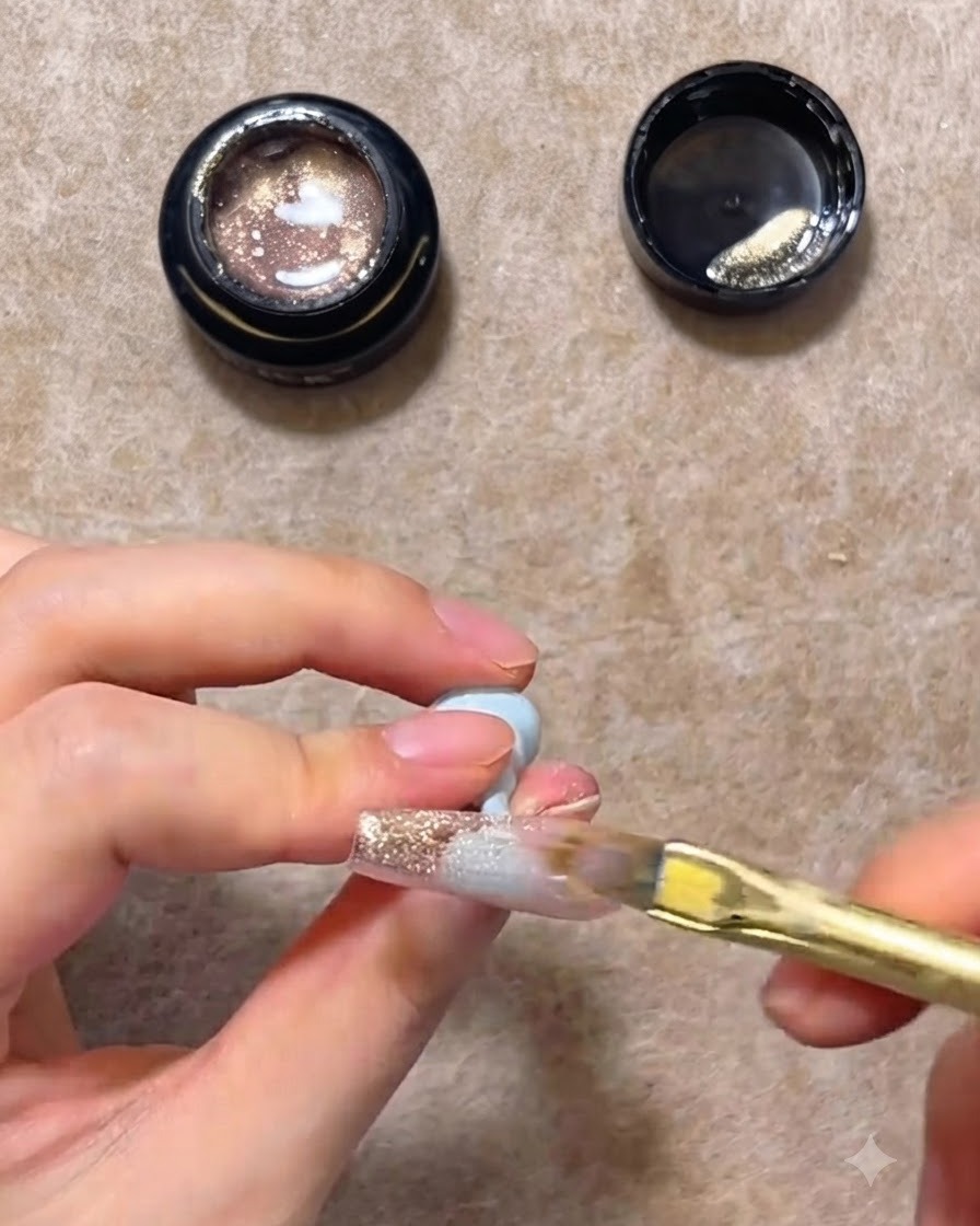 Ein flacher Nagelpinsel streicht rosa-goldenes Cat-Eye-Gel über eine transparente Nagelverlängerung nach unten, um einen Farbverlauf zu erzeugen.