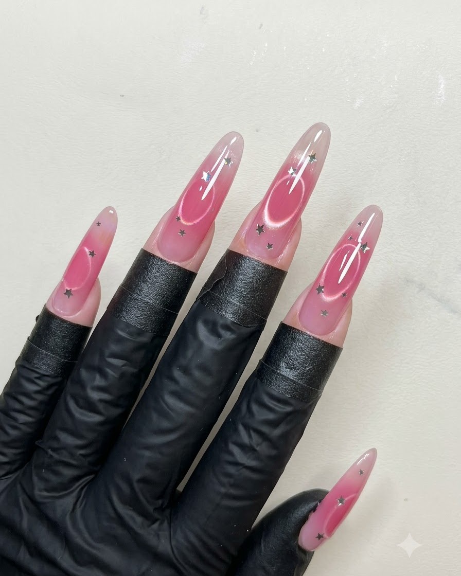 Ein Pinsel trägt eine glatte Schicht glänzenden Überlack über lange, mandelförmige Nägel auf, die mit Sternen-Pailletten und Cat-Eye-Art verziert sind.