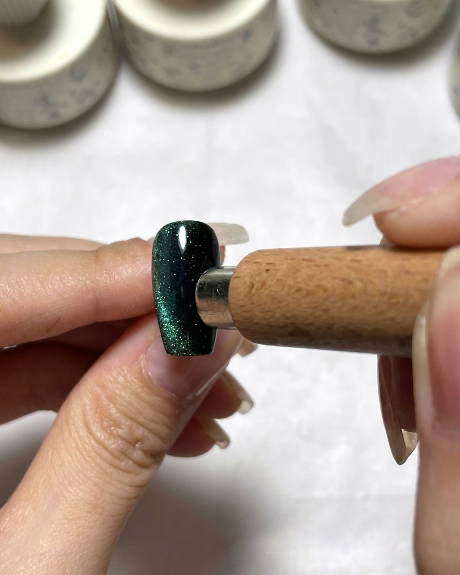 Ein Magnetwerkzeug wird neben einem dunkelgrünen Cat Eye Nagel positioniert, um das Pigment zu verteilen.