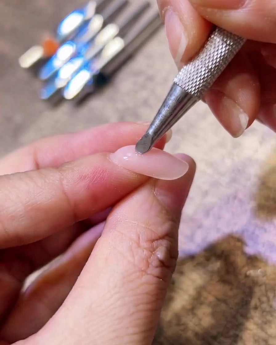 Nahaufnahme eines metallischen Applikators, der rosa Blush-Pigment in die Mitte eines Nude-Gelnagels tupft.