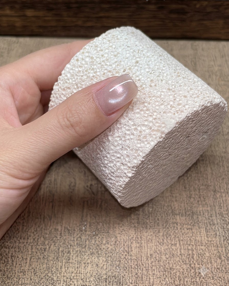 Nahaufnahme eines einzelnen Nagels vor einem strukturierten weißen Block mit einer weichen Champagner Glasperlen Cat-Eye-Reflektion.