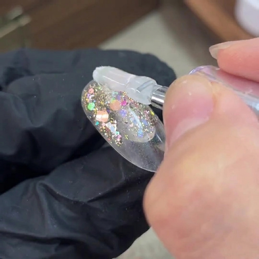 Ein Pinsel trägt klares Base-Gel über eine ausgehärtete Glitzerschicht auf einem Nagel auf.