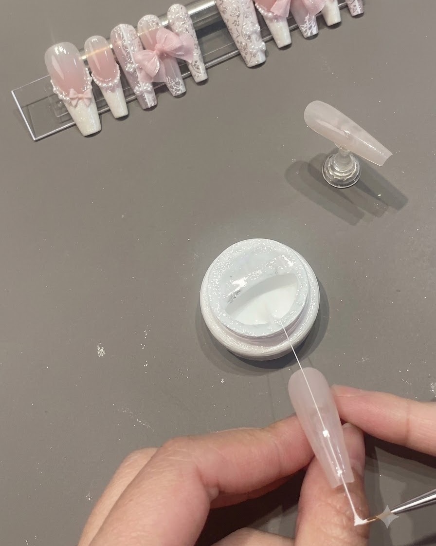 Ein schmaler Strang weißes Spider-Gel wird über der Nageloberfläche neben einem geöffneten Tiegel gezogen.