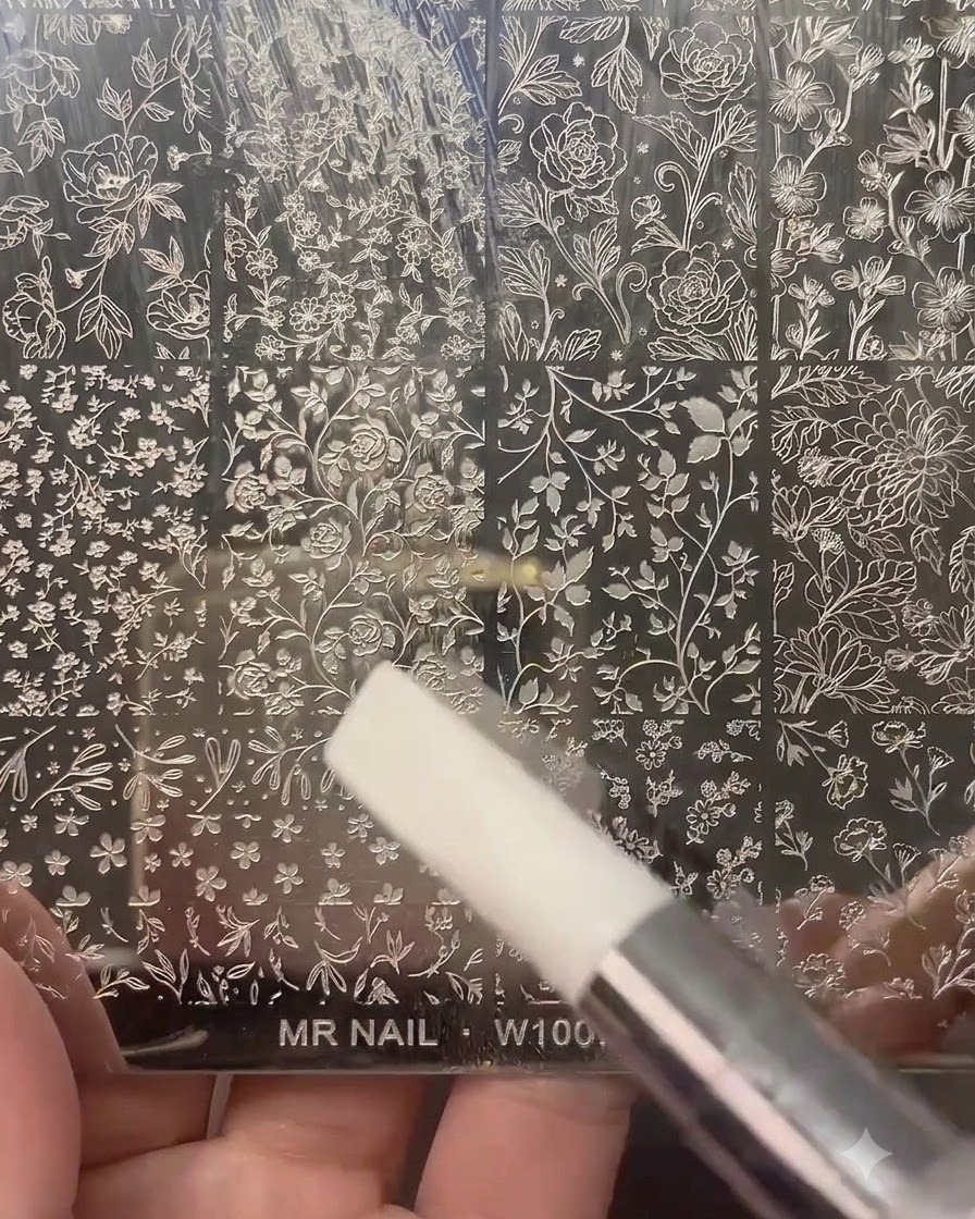Ein klarer Nagelstempel drückt auf eine gravierte Metallstempelplatte mit verschiedenen Blumen- und Botanikmustern.