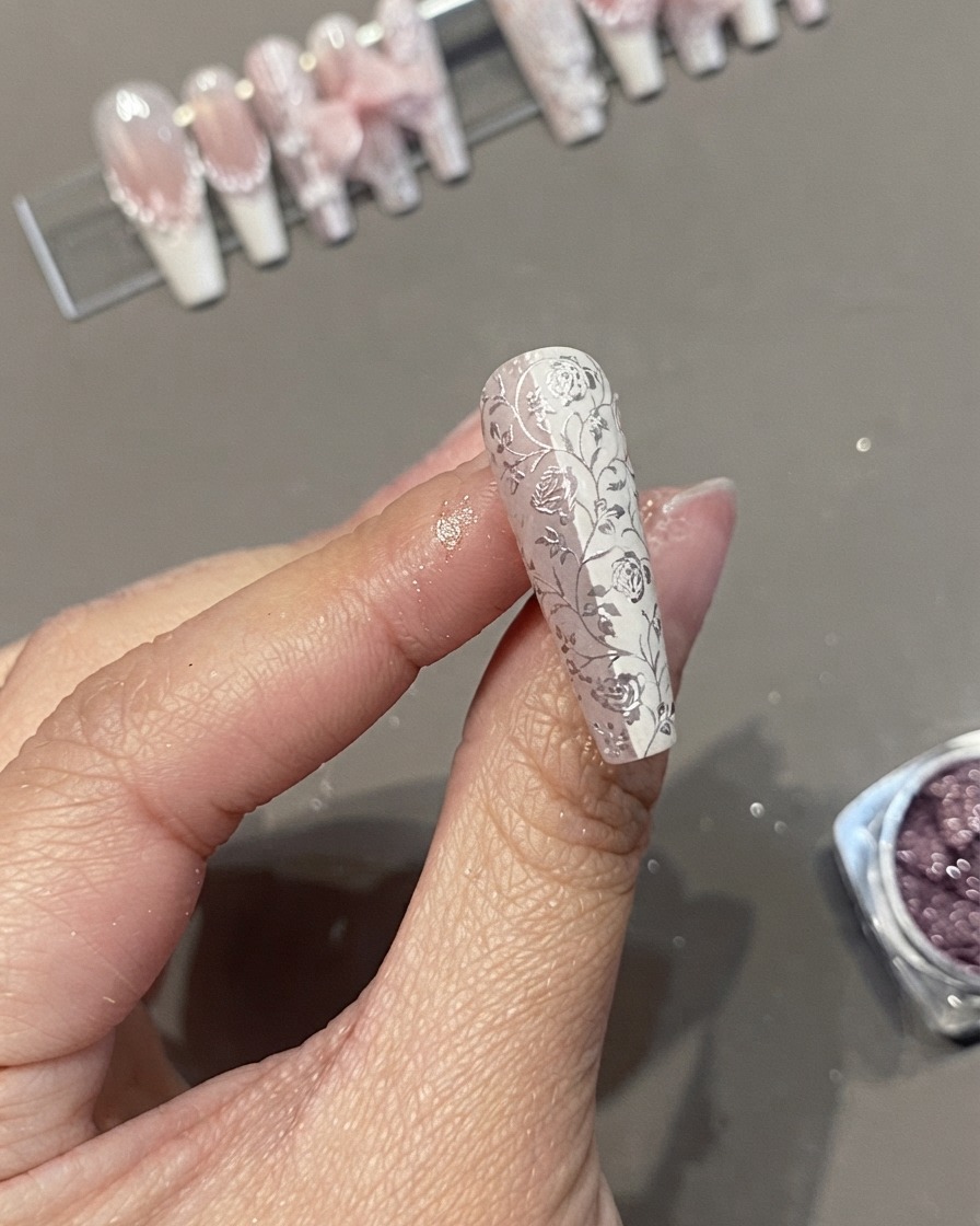 Nahaufnahme eines langen Coffin Press-on Nagels mit einem detaillierten weißen Blumen-Spitzen- und Chrommuster, gehalten zwischen Fingern.