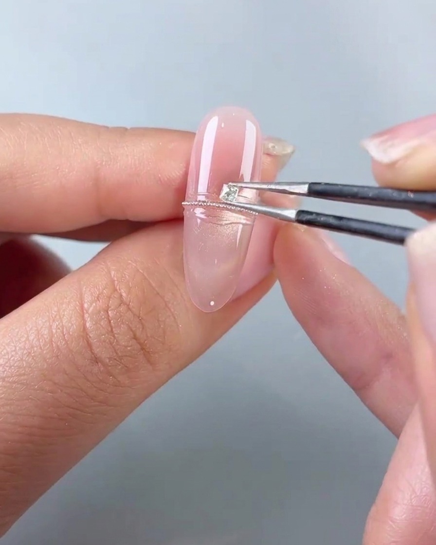 Eine Pinzette platziert einen kleinen quadratischen Diamanten neben eine Reihe von Stahlperlen auf einem rosa Gel-Nagel.